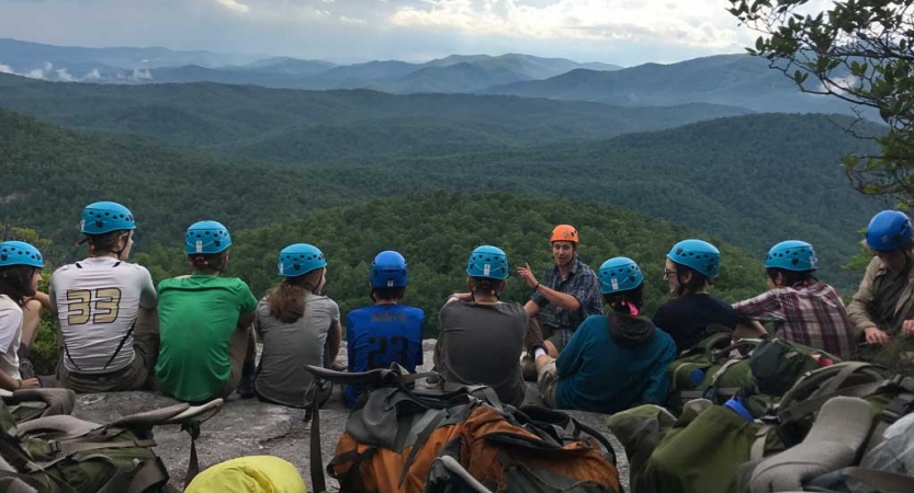 A group of students wearing helmets sit in a line and listen to an instructor speak. In the background, there is a vast and green mountainous landscape.